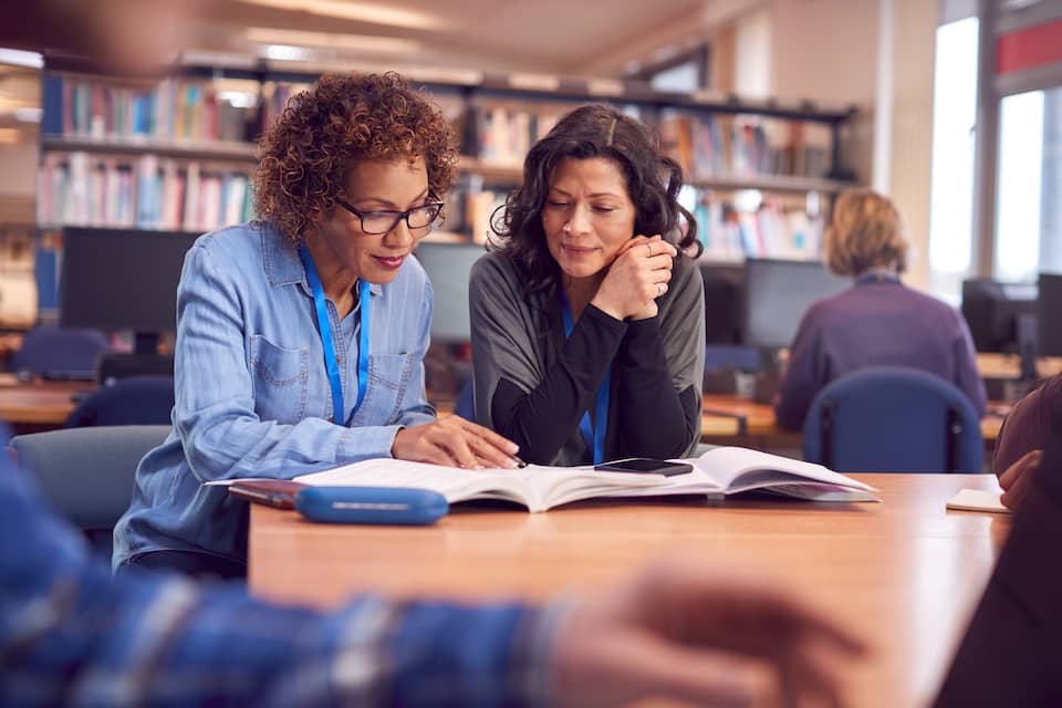 Two women look over a set of books together while seated at a table in a library.