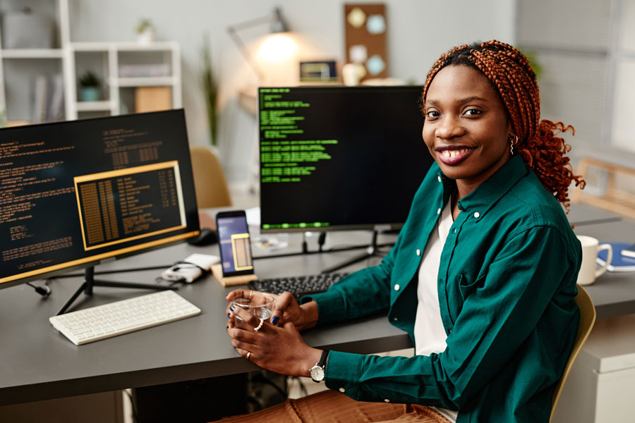 A woman smiles while computer monitors on the desk behind her depict dark screens with lines of code.